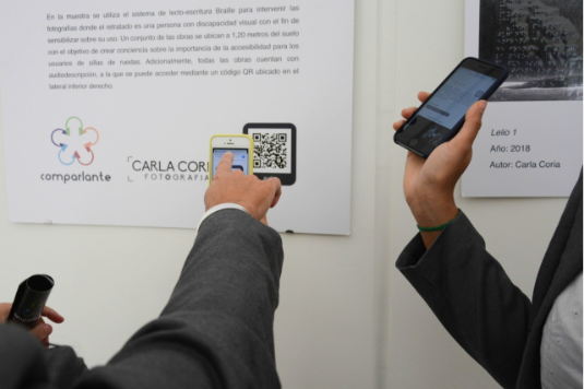 On a wall, the curatorial text of the accessible photographic exhibition “We Feel” is displayed, which includes a QR code that is being scanned by two persons with their smartphones.