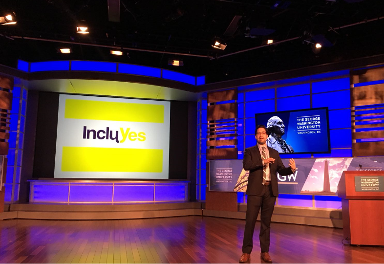 José Clautier, Institutional Relations Officer of Fundación Comparlante presenting IncluYes at the Global Public Policy Summit at George Washington University. In the background there is a screen with the IncluYes logo, and another with the University’s logo. José appears on stage addressing the audience.