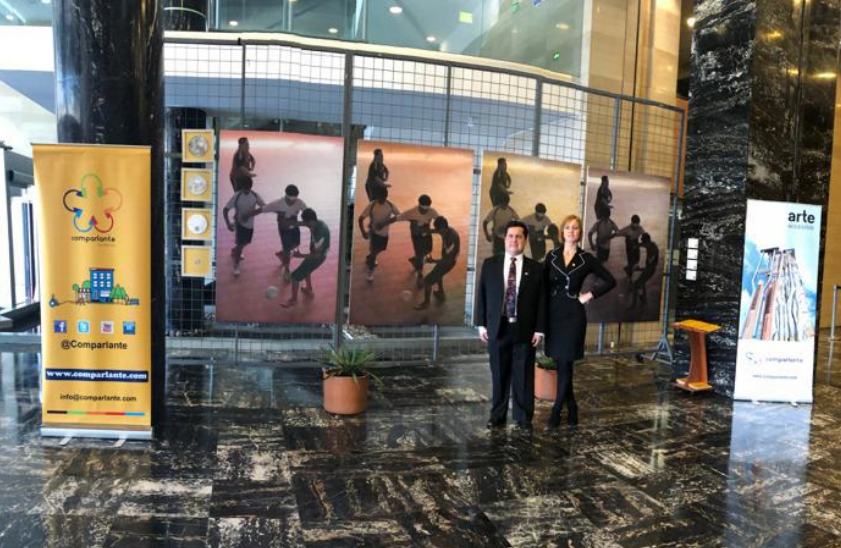 Lorena Julio and Sebastian Flores, Co-Founders of Fundación Comparlante pose with the works of the Accessible Art exhibition in the background: four photographs painted in watercolor where Lelio Sanchez is portrayed playing soccer, using different shades that simulate the way of seeing the world by persons with color blindness.