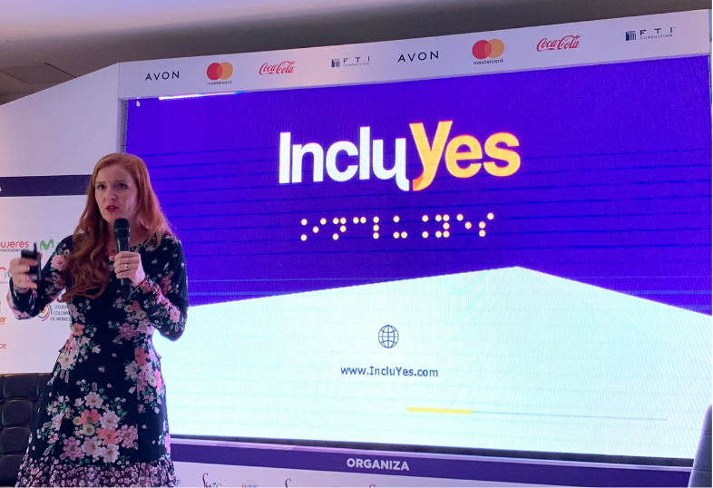 Lorena Julio, Co-Founder and President of Fundación Comparlante presenting IncluYes at the Woman Economic Forum, Cartagena 2019. In the background there is a screen with the IncluYes logo, in the center of the stage Lorena addressing the public.