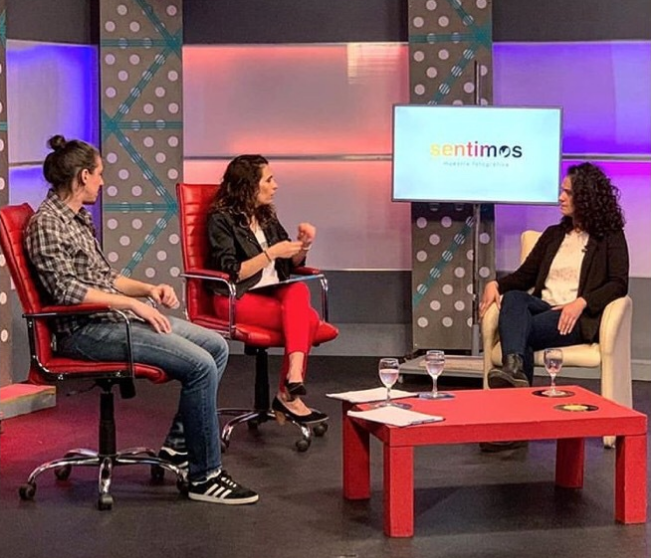 In a television studio, Carla Coria, photographer in charge of the accessible photographic exhibition “We Feel” appears next to two interviewers. They are all sitting in red chairs. In the background there is a screen with the logo of the exhibition on a white background.