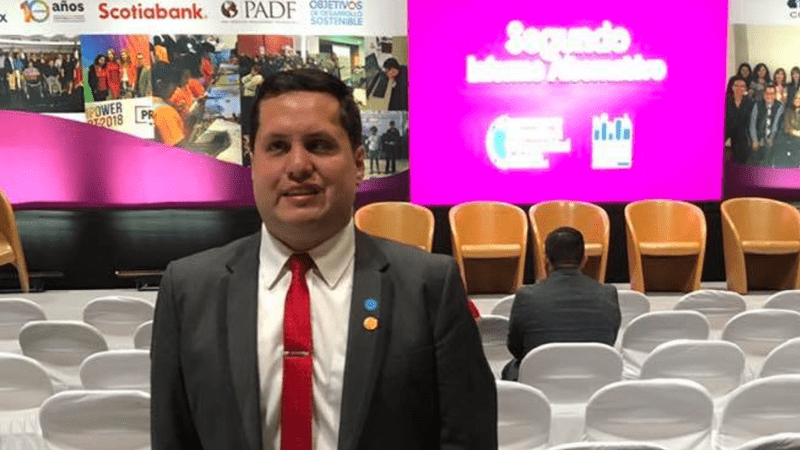 Photo of Sebastián Flores smiling. Behind him, there are several chairs and the scenario of the event with banners of the different sponsors.