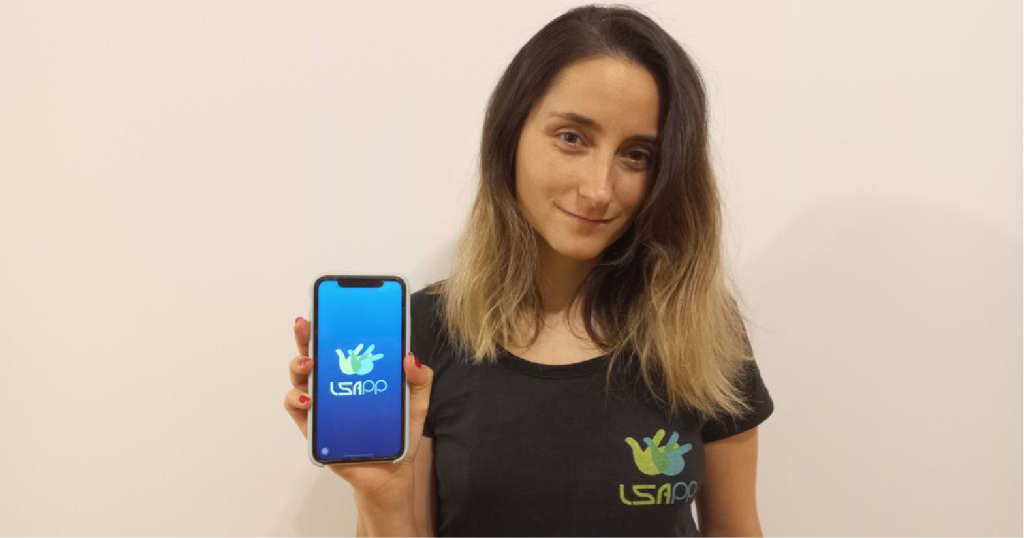 Photo of Vanesa Barán. She is wearing a t-shirt with the LSApp logo and showing her cellphone since the App can be seen in its screen.