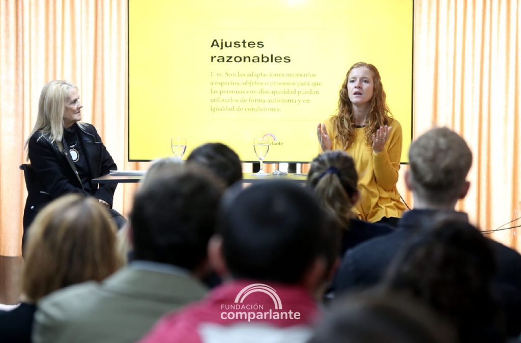 Lorena Julio, opposite the camera whilst speaking to the audience, Beside her, Ángela Bedouret seeing and carefully listening to her. In the foreground, it can be seen blurry the attendees who listen to Lorena. Behind her, a screen displaying a banner which says “Reasonable Adjustments”.