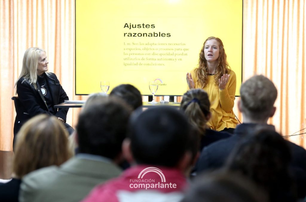 Lorena Julio, de frente a cámara mientras habla al público presente. A su lado, Ángela Bedouret mirándola y escuchando atentamente. En primer plano, desenfocados, se aprecian las espaldas de los asistentes que escuchan a Lorena. Detrás de ella, una pantalla que proyecta el cartel que dice “Ajustes Razonables”.