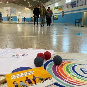 In the foreground, on the floor, there is a t-shirt of Comparlante, a copy of the Manual on Accessibility for Sports Clubs and the CEF flag. Behind, further from it, there is Horacio with two members of the CEF whilst smiling at the camera.