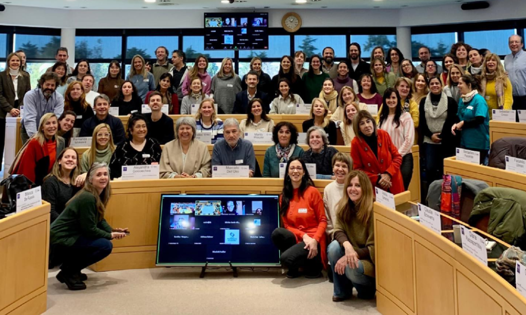Group photo with the attendees smiling at the camera. Everybody is gathered together in the center of the amphitheater next to a screen in which are displayed the attendees who connected to the meeting online.