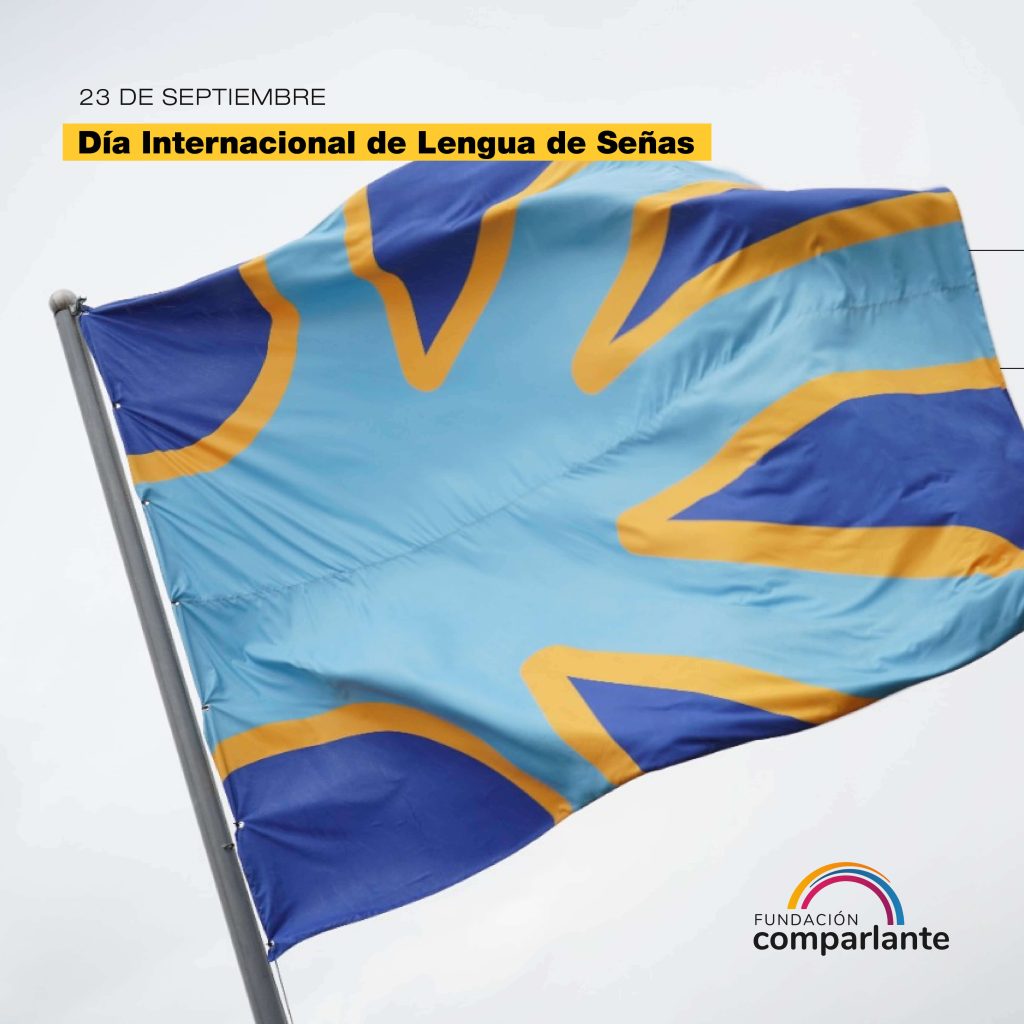 Fotografía titulada “23 de septiembre, Día Internacional de la Lengua de Señas.” Debajo, en primer plano, la bandera de la comunidad sorda flameando en un mástil. La bandera es de un lienzo azul marino y por encima, ocupando el centro y gran parte de la superficie de la misma, se encuentra una figura de mano extendida. Esta mano es de color azul claro con bordes amarillos. Como pie de imagen, el logo de Fundación Comparlante.