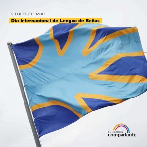 Fotografía titulada “23 de septiembre, Día Internacional de la Lengua de Señas.” Debajo, en primer plano, la bandera de la comunidad sorda flameando en un mástil. La bandera es de un lienzo azul marino y por encima, ocupando el centro y gran parte de la superficie de la misma, se encuentra una figura de mano extendida. Esta mano es de color azul claro con bordes amarillos. Como pie de imagen, el logo de Fundación Comparlante.