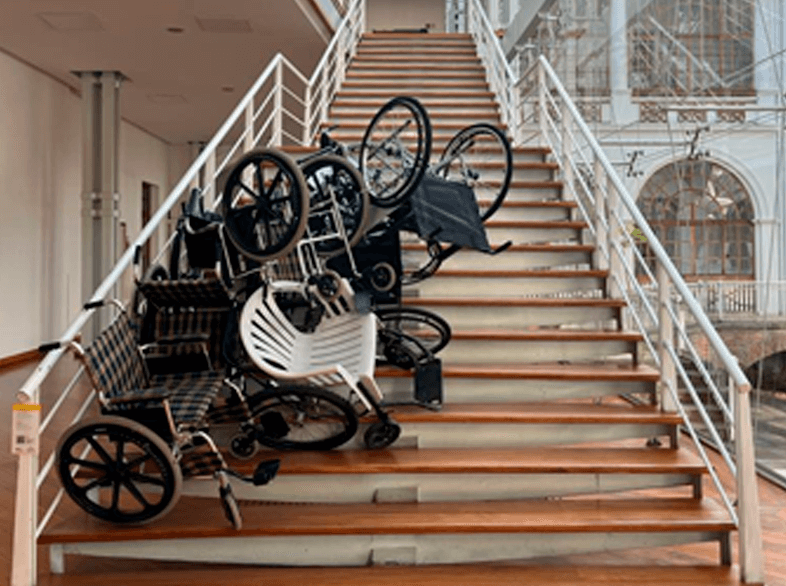 Instalación de un conjunto de 10 sillas de ruedas encimadas obstruyendo una sección de la escalera central del edificio.